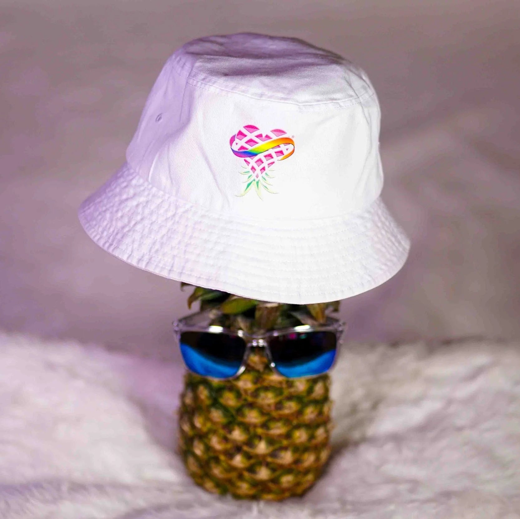 Pineapple wearing sunglasses and a white bucket hat with a colorful logo on a soft surface.